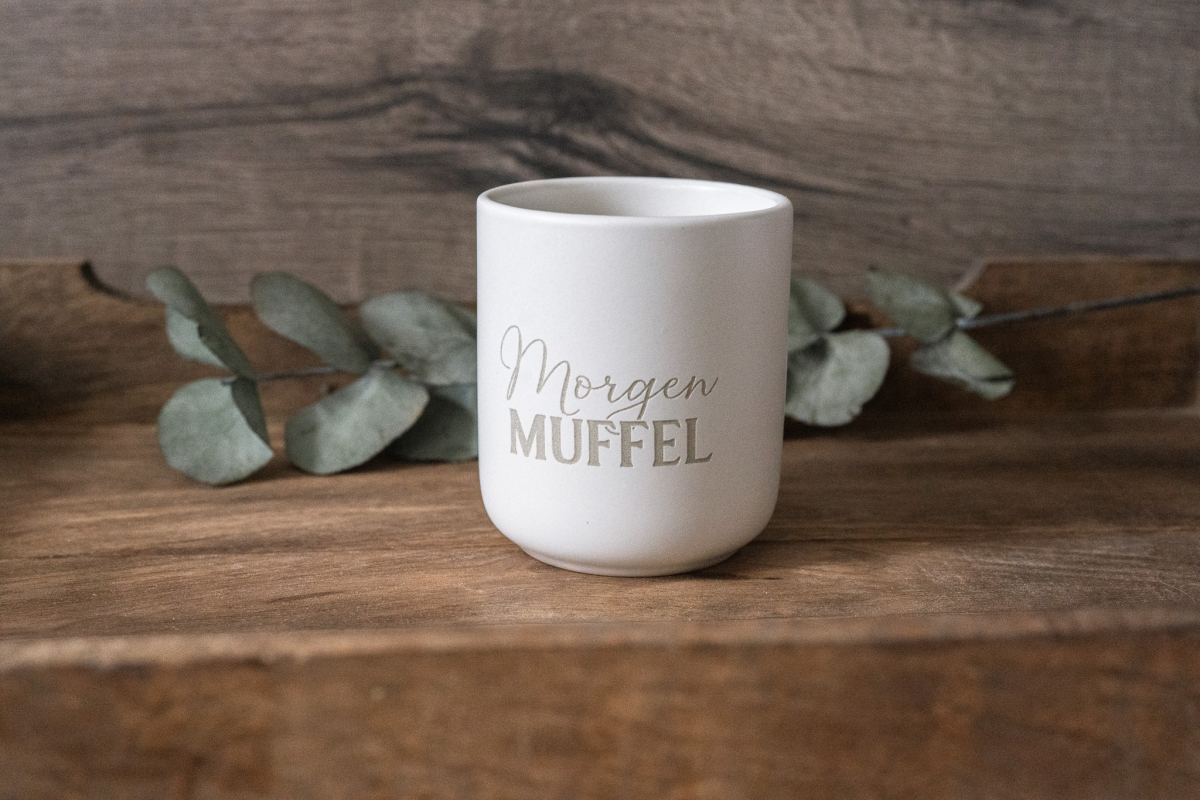 Matte weiße Keramiktasse mit Gravur „Morgen MUFFEL“ auf Holzuntergrund, dekoriert mit Eukalyptus.